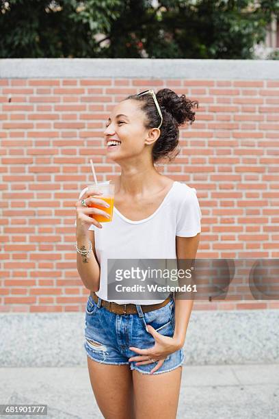 happy young woman holding orange juice outdoors - women in daisy dukes stock-fotos und bilder