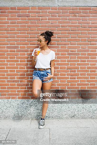 young woman leaning against a brick wall drinking orange juice - denim shorts stock pictures, royalty-free photos & images