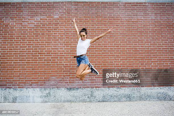 exuberant young woman jumping in front of a brick wall - solo una donna giovane foto e immagini stock