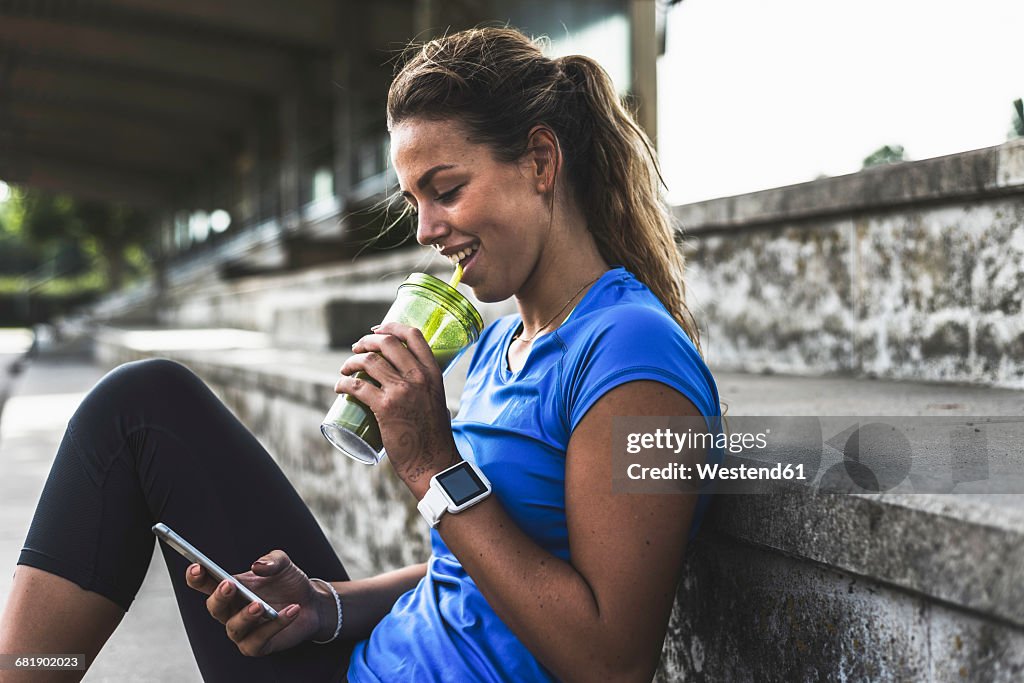Sportive young woman sitting on grandstand with cell phone and drinking mug