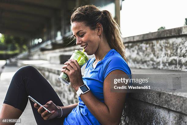 sportive young woman sitting on grandstand with cell phone and drinking mug - drinking smoothie stock pictures, royalty-free photos & images