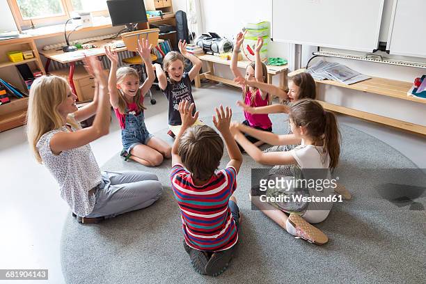 teacher and pupils sitting in a circle on floor of their classroom raising hands - bambini seduti in cerchio foto e immagini stock