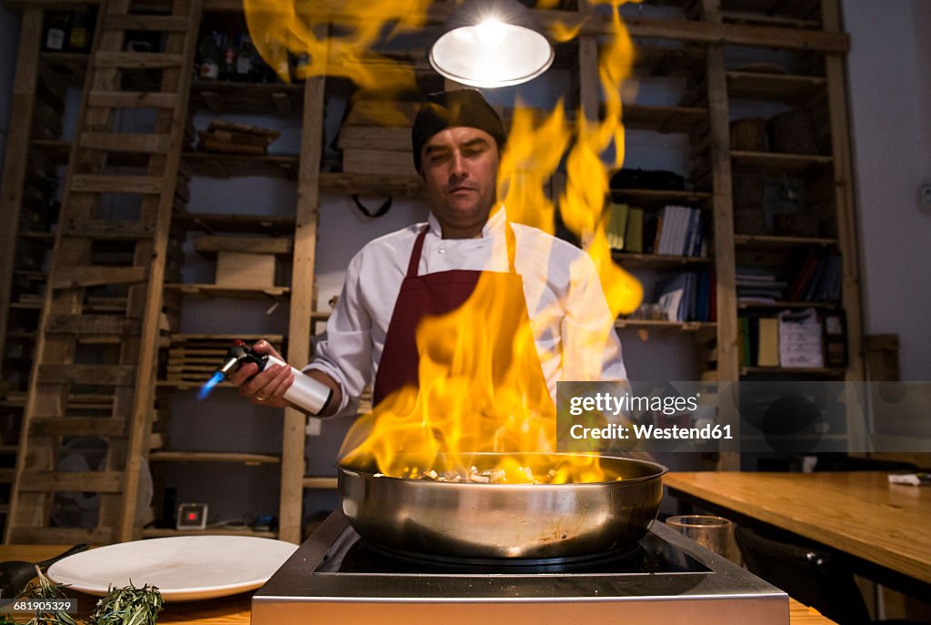 Man flambing beef cheeks with sauteed vegetables in a pan using a kitchen torch