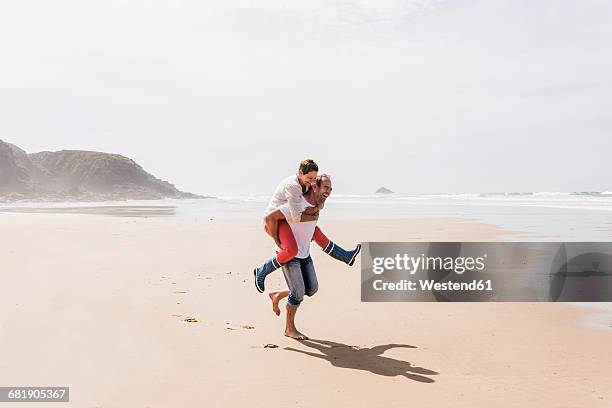 happy mature man carrying wife piggyback on the beach - couple et vacances photos et images de collection