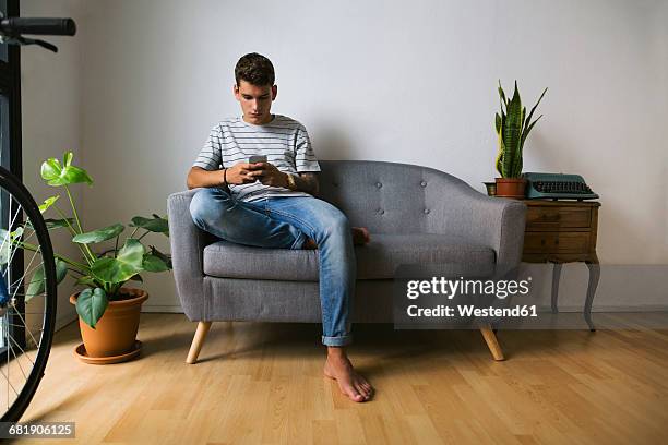 teenage boy sitting on couch at home looking at cell phone - teen boy barefoot stock pictures, royalty-free photos & images