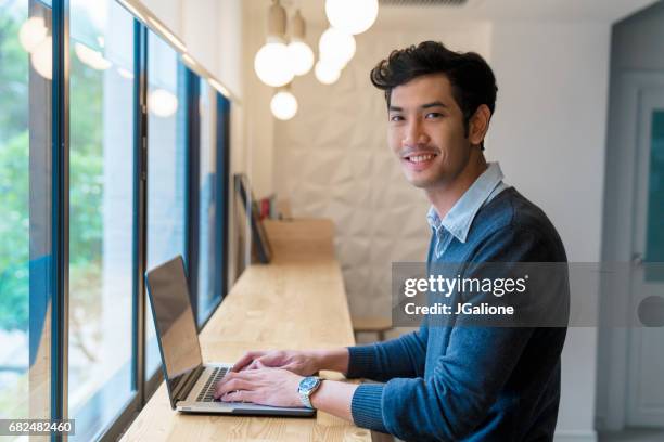 retrato de un varón adulto joven sentado con su ordenador portátil en una oficina moderna - thai ethnicity fotografías e imágenes de stock