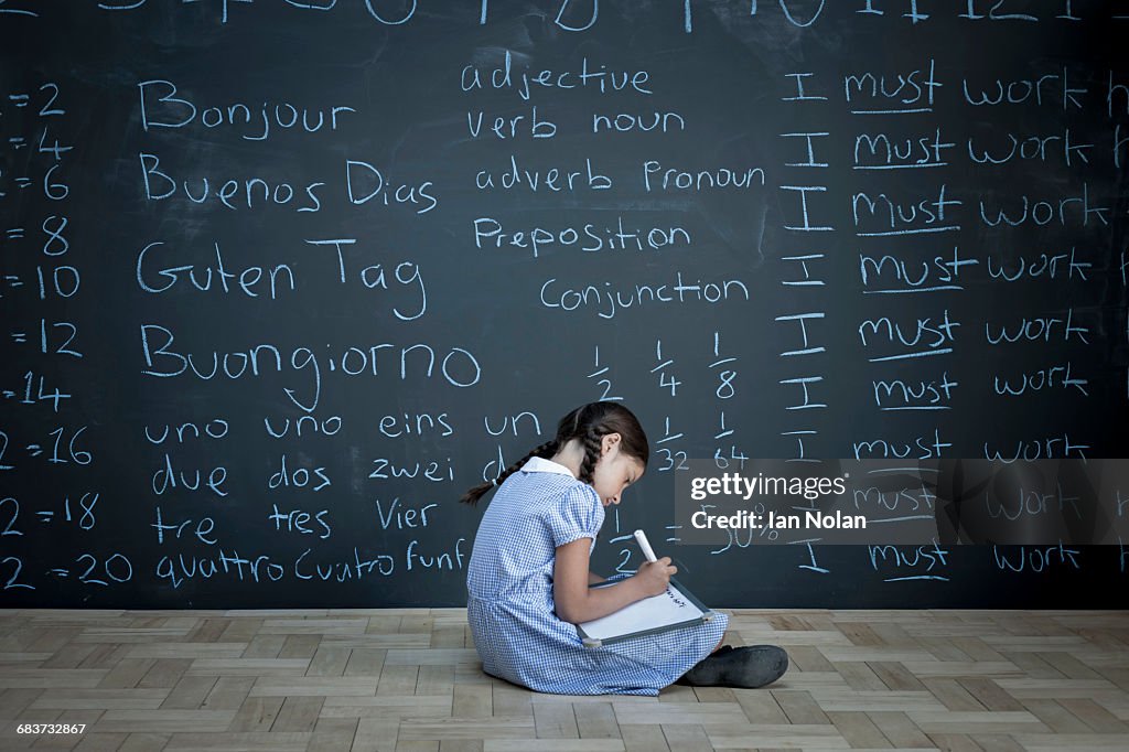 Schoolgirl sitting studying in front of large chalkboard with schoolwork chalked on it