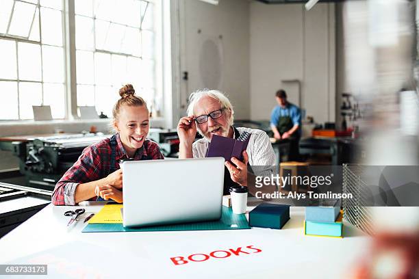 senior male craftsman laughing and looking at laptop with young woman in book arts workshop - craft show stock pictures, royalty-free photos & images
