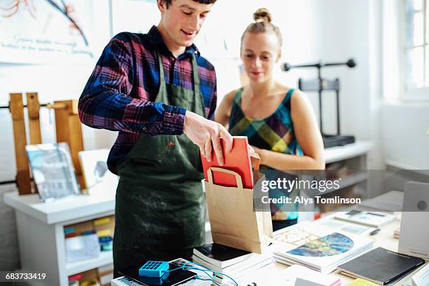 young craftsman with customer, placing book into bag in arts bookshop - sold engelskt begrepp bildbanksfoton och bilder