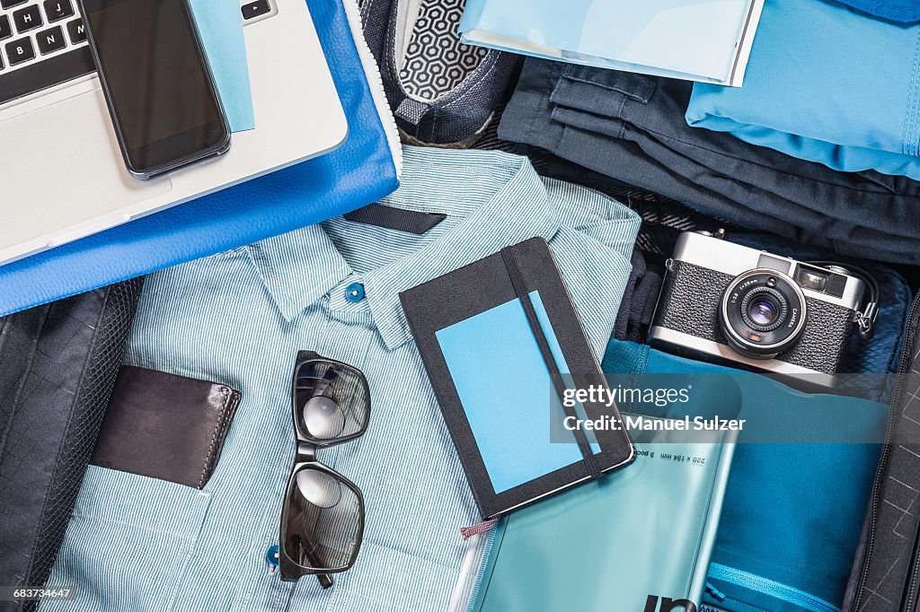 Overhead detail of packed suitcase with blue shirt, retro camera, laptop, smartphone and notebook