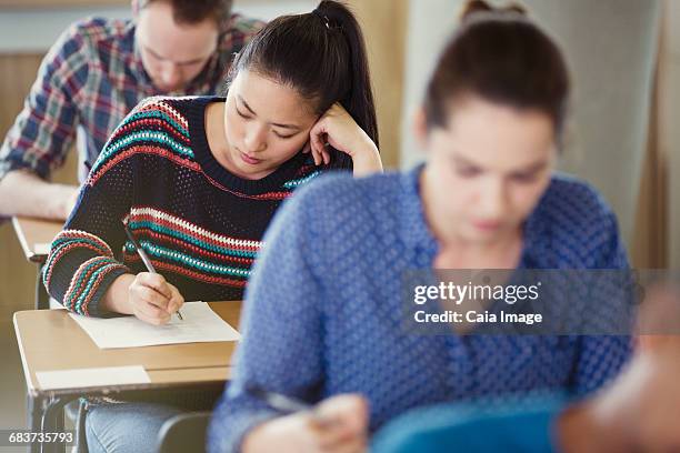 college students taking test at desks in classroom - tentamenzaal stockfoto's en -beelden