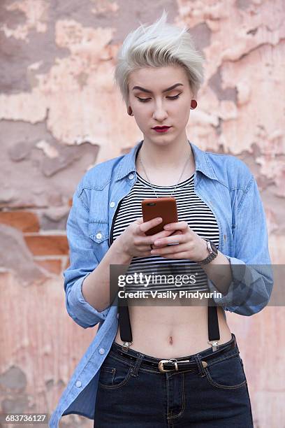 beautiful woman using phone while standing against old wall - women in suspenders stock pictures, royalty-free photos & images