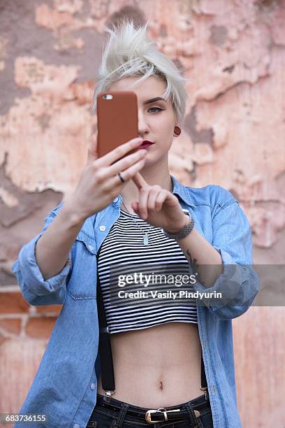 confident young woman taking selfie through smart phone while standing against wall - women in suspenders stock pictures, royalty-free photos & images