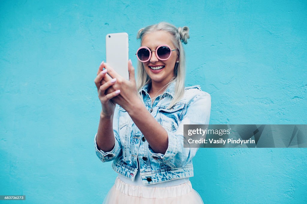 Happy fashionable young woman talking selfie against blue wall