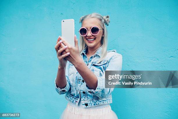 happy fashionable young woman talking selfie against blue wall - blonde woman selfie foto e immagini stock