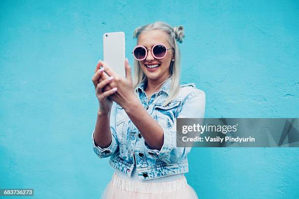 happy fashionable young woman talking selfie against blue wall - blonde woman selfie stock pictures, royalty-free photos & images