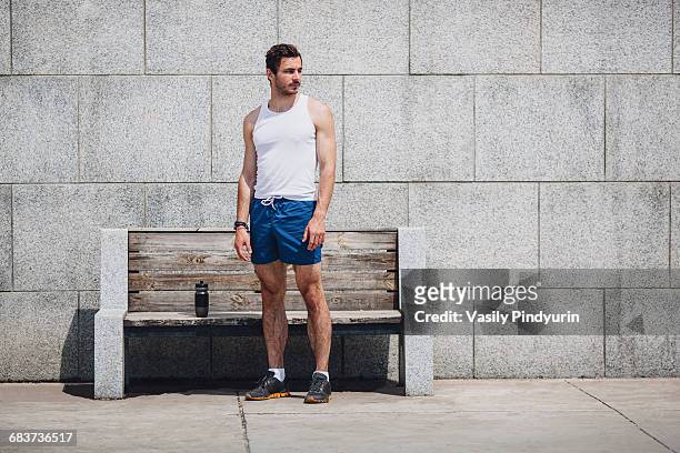 serious sportsman standing by wooden bench on sidewalk against wall - men shorts stock pictures, royalty-free photos & images