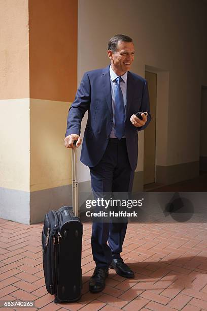 happy businessman holding mobile phone and suitcase while standing at office - costume complet photos et images de collection