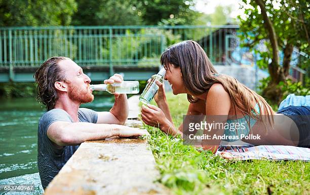 young man in river, young woman lying on grass at edge of river, man drinking from beer bottle - andreas pollok stock-fotos und bilder