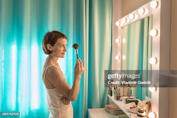 woman holding make-up brush standing in front of mirror at dressing table - dressing room stock-fotos und bilder