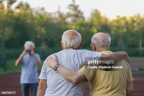 rear view of friends standing with arm around while man photographing in background - arm around friend back stock pictures, royalty-free photos & images