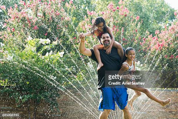 mixed race father holding daughters in backyard sprinkler - swimsuit models girls stock pictures, royalty-free photos & images