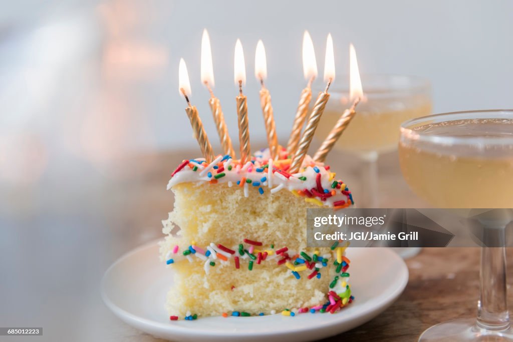 Candles burning on slice of cake with sprinkles near champagne