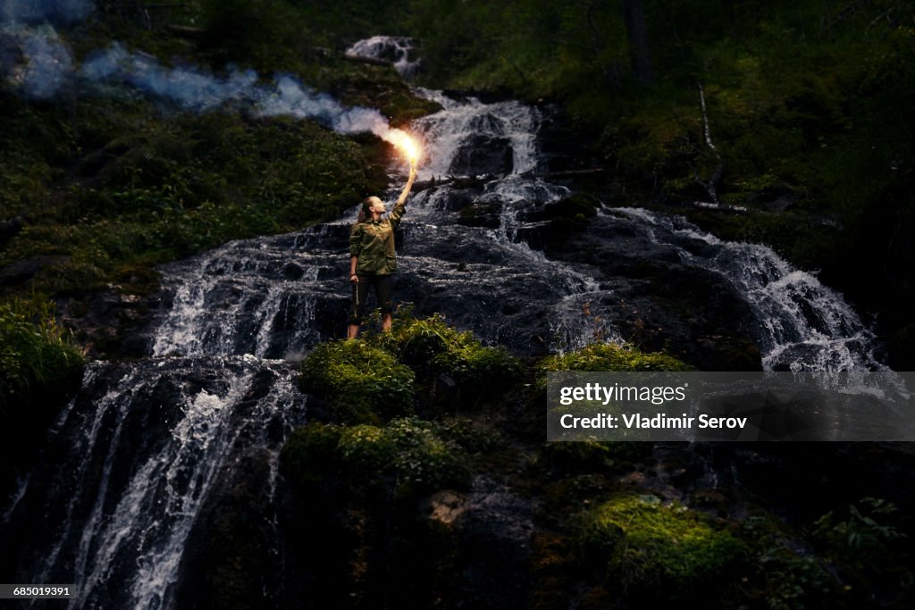 Caucasian girl holding smoke flare near waterfall