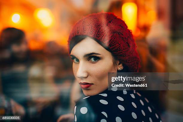 portrait of young woman wearing red hat looking through window of a pub in the evening - verführung stock-fotos und bilder