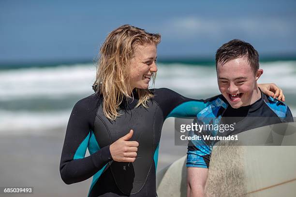 happy teenage boy with down syndrome and woman with surfboard on beach - down's syndrome fotografías e imágenes de stock