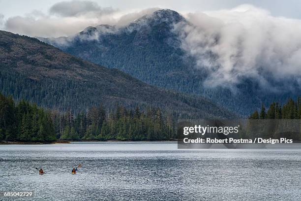 two kayakers paddling toward the mainland while clouds flow over the coastal mountains - queen charlotte islands stock pictures, royalty-free photos & images