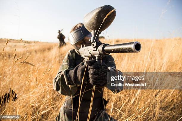 female paintball player in grass aiming with paintball gun during a paintball game - paintball stock pictures, royalty-free photos & images