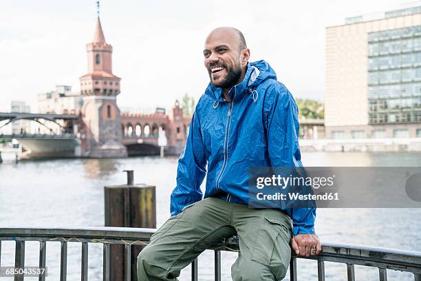 germany, berlin, laughing man wearing blue rainjacket sitting on railing in front of oberbaum bridge - oberbaumbrücke fotografías e imágenes de stock