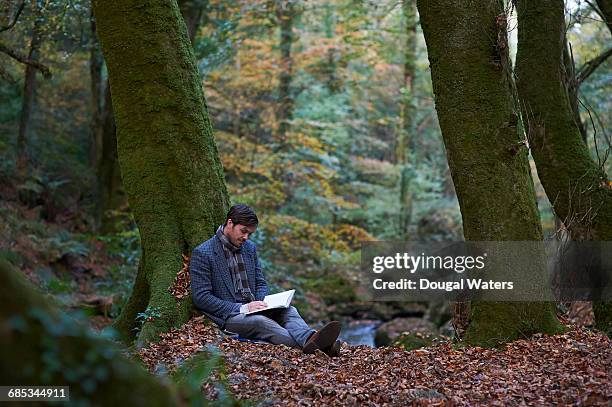 man sitting in autumn woodland reading book. - escapism reading stock pictures, royalty-free photos & images