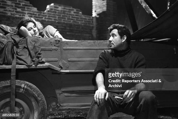 Italian actress Stefania Sandrelli sleeping inside the cargo bed of a van. Beside her, sitting on the running board of the vehicle and looking at...