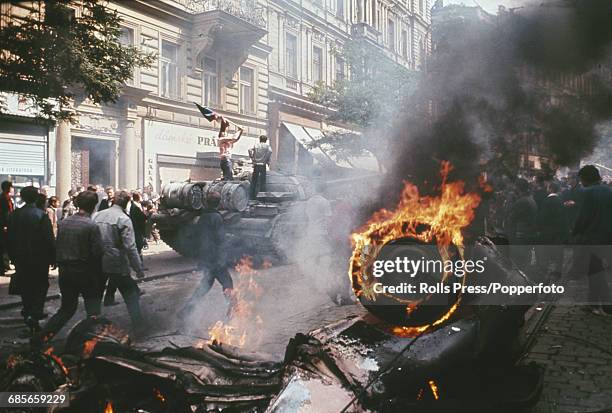 View of protesters and residents of Prague, Czechoslovakia standing on and around a Russian tank as a wrecked motor vehicle burns on a city street in...