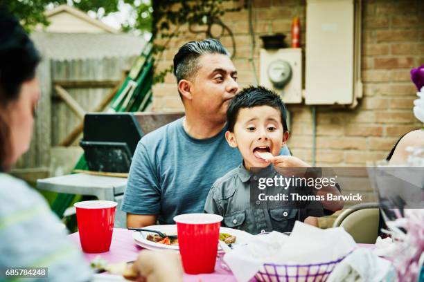 young boy sitting on fathers lap during family birthday party eating tortilla chip - tortilla flatbread stock pictures, royalty-free photos & images