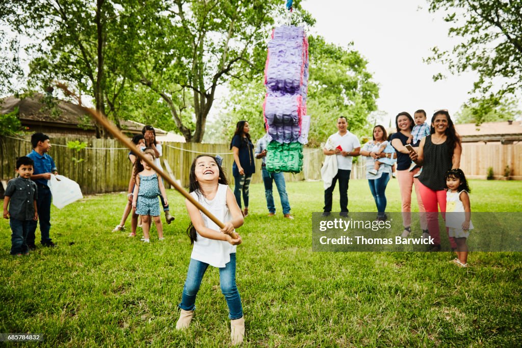 Laughing young girl trying to break open pinata during family birthday party in backyard