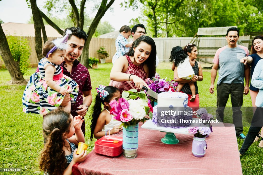 Mother cutting cake for daughter during first birthday party with family in backyard