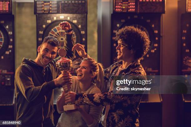group of happy friends with winning trophy celebrating victory in dartboard game. - dart stock pictures, royalty-free photos & images