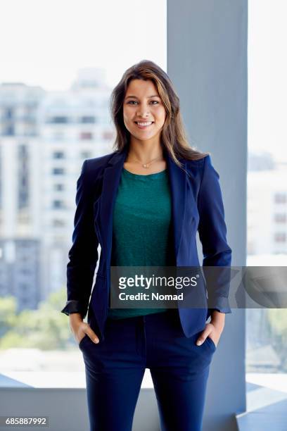 portrait of attractive female executive standing in creative office. smiling businesswoman is with hands in pockets against window. she is wearing smart casuals. - blue blazer stock pictures, royalty-free photos & images