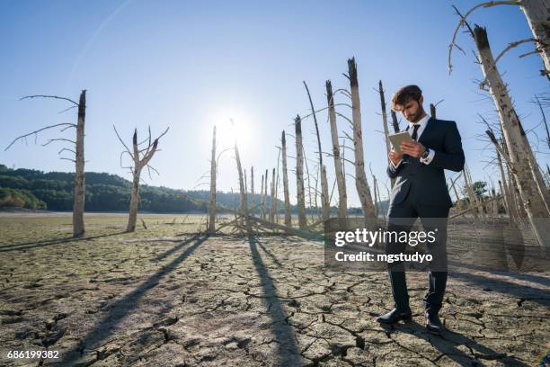 businessman with digital tablet working on cracked earth - climate change finance stock pictures, royalty-free photos & images