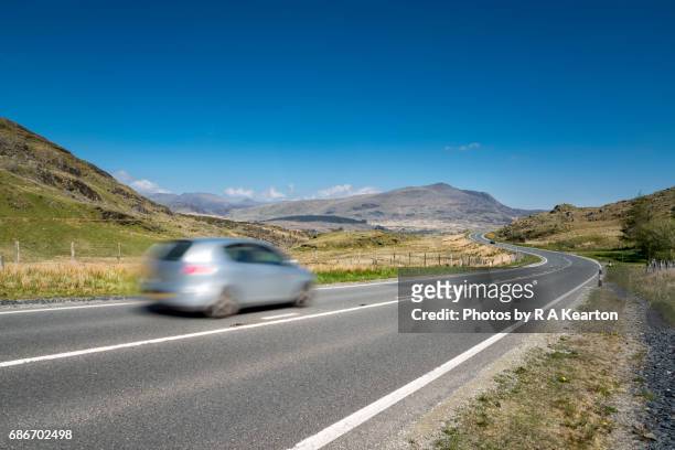 silver car speeding along a road in snowdonia, north wales, uk - snowdonia wales stock pictures, royalty-free photos & images