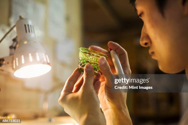 a craftsman at work in a glass makers studio workshop, inspecting a small glass bowl against a beam of light, holding a pen.  - kanto region foto e immagini stock