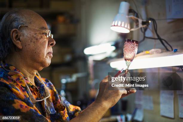 a senior craftsman at work in a glass makers studio workshop, in inspecting red wine glass with cut glass decoration against the light.  - 工芸品 ストックフォトと画像