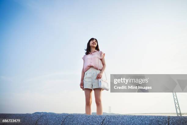 young woman in shorts and pink shirt standing alone in open space, with a large suspension bridge in the background - mirar alrededor fotografías e imágenes de stock