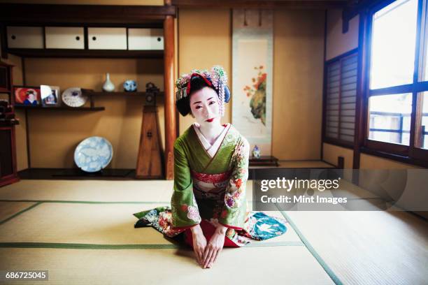 a woman dressed in the traditional geisha style, wearing a kimono and obi, with an elaborate hairstyle and floral hair clips, with white face makeup with bright red lips and dark eyes, kneeling in a traditional pose.  - geisha in training stock pictures, royalty-free photos & images