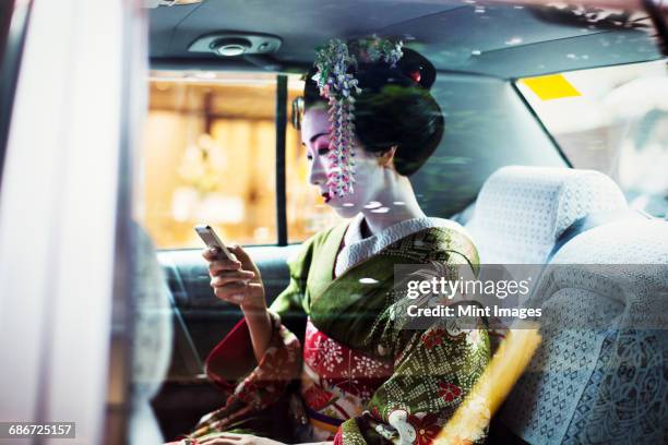 a woman dressed in the traditional geisha style, wearing a kimono and obi, with an elaborate hairstyle and floral hair clips, with white face makeup with bright red lips and dark eyes in a car using a smart phone.  - geisha in training stock pictures, royalty-free photos & images