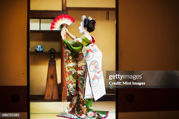 a woman dressed in the traditional geisha style, wearing a kimono and obi, with an elaborate hairstyle and floral hair clips, with white face makeup with bright red lips and dark eyes. standing in a classic pose with fan raised, side view.  - geisha in training stock pictures, royalty-free photos & images