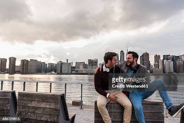 usa, new york city, two happy young men with headphones and cell phone sitting at east river - panorama nyc day 2 foto e immagini stock